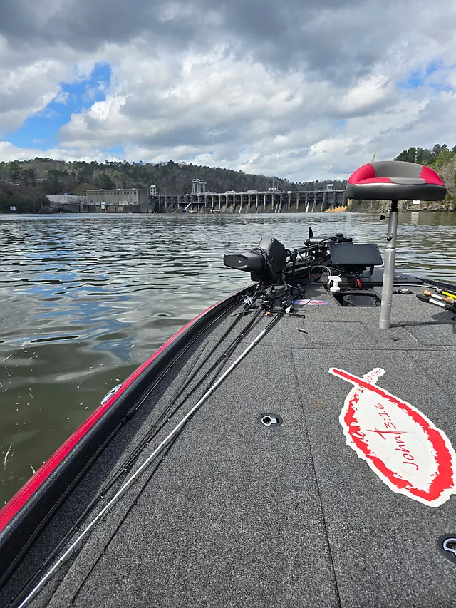 The bow of a red bass boat with rods laid out and a John 3:16 decal on the carpet, idling below a dam